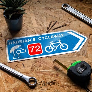 May include: A distressed blue and white sign for Hadrian's Cycleway, featuring a bicycle symbol and the number 72. The image includes tools like a wrench, screws, and a tape measure, alongside a small potted plant.