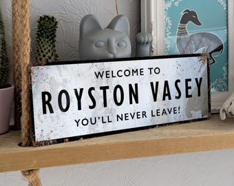 May include: A rectangular metal sign with the text "WELCOME TO ROYSTON VASEY YOU'LL NEVER LEAVE!" in black letters. The sign has a distressed, aged appearance with a white background and black border. The sign is displayed on a wooden shelf.