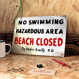 May include: A weathered metal sign with the words "NO SWIMMING HAZARDOUS AREA BEACH CLOSED" in black and red. The sign is on a wooden surface, with a white mug, a red pen, and a small card that says "THANK YOU!" nearby.