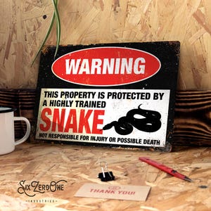 May include: A black and red metal sign with the text "WARNING" in a red circle. The sign reads "This property is protected by a highly trained snake. Not responsible for injury or possible death."