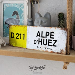 May include: A yellow and white metal sign with black text that reads "D 211 ALPE D'HUEZ ALT. 1860m".