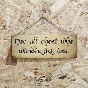 May include: A rustic wooden sign with a distressed, aged look. The sign features the quote "Not all those who wander are lost" in a stylized, black font. The sign is hanging from a string.