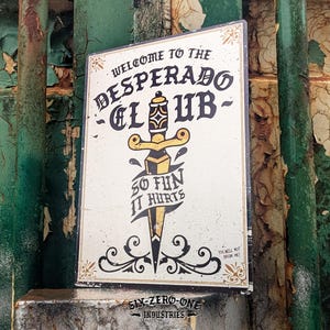 May include: A rectangular metal sign with a cream background featuring a black and gold dagger illustration. The text reads: WELCOME TO THE DESPERADO CLUB, SO FUN IT HURTS, and YOU WILL NOT BREAK ME. The bottom includes the text SIX-ZERO-ONE INDUSTRIES.