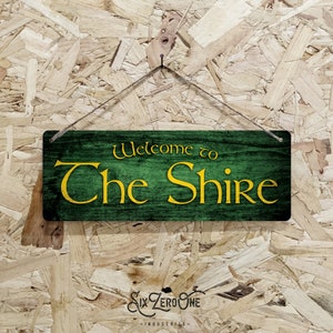 May include: A wooden sign with green paint and a yellow inscription that reads "Welcome to The Shire".
