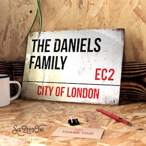 May include: White metal sign with black text that reads "The Daniels Family" and red text that reads "City of London EC2".