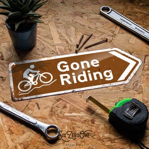 May include: A brown metal sign with the words "Gone Riding" and a bicycle graphic. The sign is surrounded by tools, including a spanner, tape measure, and screws. A small potted plant is also present.