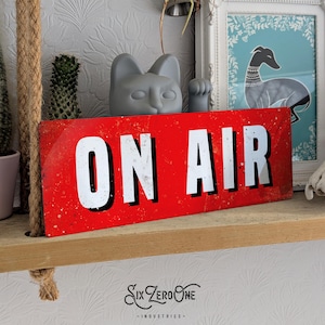 May include: A rectangular red metal sign with the words "ON AIR" in large white block letters. The sign has a distressed, vintage appearance. The text "Six Zero One - Industries" is below the sign.