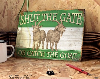 Puede incluir: Un letrero metálico desgastado con el texto "SHUT THE GATE OR CATCH THE GOAT" en letras blancas. Presenta dos ilustraciones detalladas de cabras sobre un fondo verde y blanco. El letrero está sobre una superficie de madera.