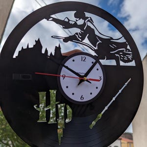 Op de afbeelding: Een zwarte vinylplaatklok met een silhouet van een persoon op een bezem, een kasteel en een toverstaf. De klok heeft een witte wijzerplaat met zwarte cijfers en wijzers. De letters "HP" zijn ook zichtbaar.