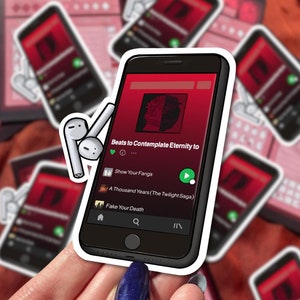 Puede incluir: Una pegatina blanca con un contorno negro de un tel&eacute;fono inteligente reproduciendo m&uacute;sica. La pantalla muestra el texto "Beats to Contemplate Eternity to" y una lista de canciones: "Show Your Fangs", "A Thousand Years (The Twilight Saga)" y "Fake Your Death".