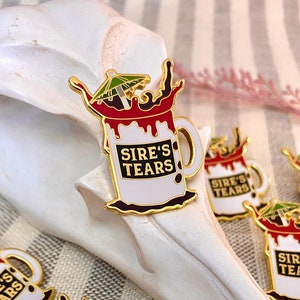 Op de afbeelding: Gouden emaille pin met een witte koffiemok met rood vloeistof dat eruit spuit. De mok heeft een zwarte omtrek en de tekst "Sire's Tears" in zwarte letters. Een kleine groene paraplu bevindt zich bovenop de mok.