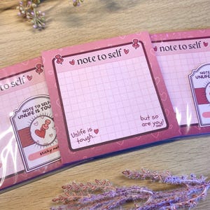 Puede incluir: Blocs de notas adhesivas rosas con el texto "note to self" y "Unlife is tough... but so are you!" impreso. Cada bloc tiene un patrón de cuadrícula e ilustraciones de corazones. Los blocs están empaquetados en plástico transparente.