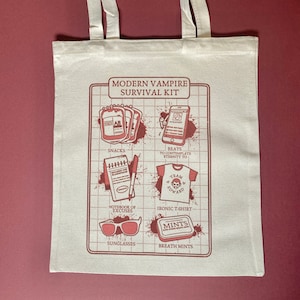 Può includere: Una borsa di tela bianca con un design grafico rosso che dice "Modern Vampire Survival Kit" e include illustrazioni di sacche di sangue, snack, un quaderno, occhiali da sole, mentine per l'alito, una maglietta e un telefono.