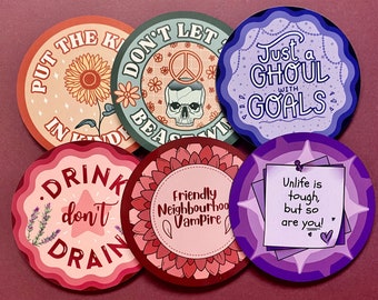 May include: Six round coasters with different designs and sayings. The coasters are pink, purple, red, and green. The sayings include "Put the Kindness in", "Don't Let the Beast Win", "Just a Ghoul with Goals", "Drink Don't Drain", "Friendly Neighbourhood Vampire", and "Unlife is tough, but so are you!"