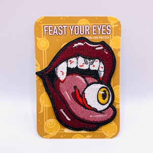 Peut inclure: Un patch thermocollant brodé rouge avec une grande bouche aux crocs et une langue qui sort, tenant un globe oculaire blanc avec un iris jaune. Le patch indique "Feast Your Eyes" et "Iron-On Patch".