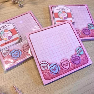 Puede incluir: Notas adhesivas rosas con un patrón de cuadrícula y el texto "UNDEAD LOVE HEARTS". Las notas presentan diseños de corazones de caramelo con frases como "HAUNT ME" y "BE MY BOO".