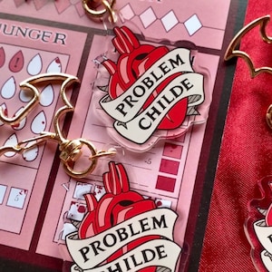 Pode incluir: Dois pingentes de acrílico transparente com um coração vermelho e uma faixa branca que diz "Problem Childe". Os pingentes estão presos a um chaveiro de metal dourado.