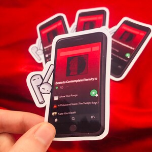 Puede incluir: Una pegatina de un tel&eacute;fono inteligente reproduciendo m&uacute;sica en un servicio de transmisi&oacute;n. La pantalla del tel&eacute;fono muestra la canci&oacute;n "Beats to Contemplate Eternity to" del artista "Show Your Fangs". La pegatina tambi&eacute;n incluye un par de auriculares inal&aacute;mbricos blancos.
