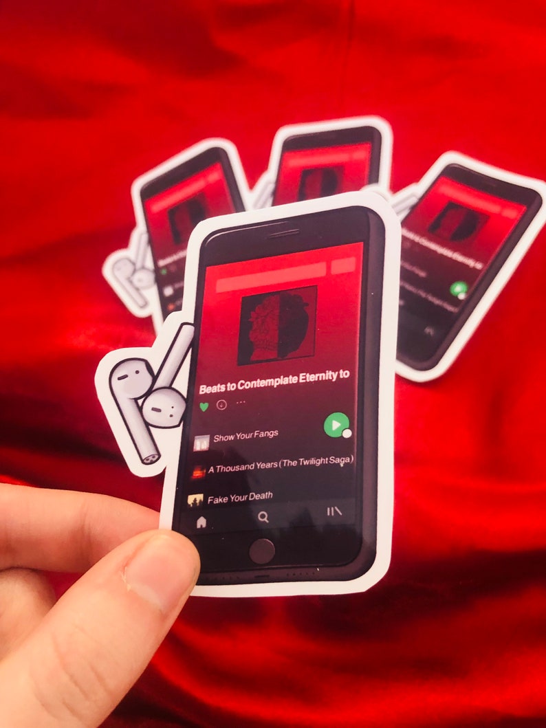 Puede incluir: Una pegatina de un tel&eacute;fono inteligente reproduciendo m&uacute;sica en un servicio de transmisi&oacute;n. La pantalla del tel&eacute;fono muestra la canci&oacute;n "Beats to Contemplate Eternity to" del artista "Show Your Fangs". La pegatina tambi&eacute;n incluye un par de auriculares inal&aacute;mbricos blancos.