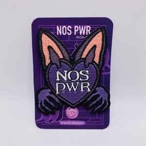 May include: Purple iron-on patch with a bat holding a heart that says "NOS PWR" in white text. The patch is on a purple card with the text "NOS PWR" and "@enthralledyet" on it.