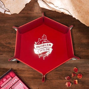 May include: A hexagonal red dice tray with silver snap fasteners at the corners. The centre features a white graphic of an anatomical heart with a banner containing the text "PROBLEM CHILDE" in a bold, capitalised, sans-serif typeface.