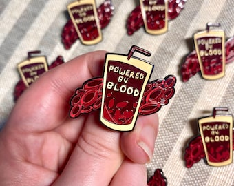 May include: A red enamel pin with a cartoon image of a glass of blood with a straw and the text "Powered by Blood". The glass is surrounded by red blood cells.