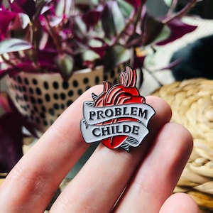 Può includere: Un cuore anatomico rosso con uno striscione bianco che recita "PROBLEM CHILDE". La spilla è un oggetto di novità.