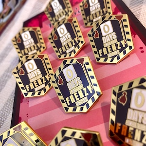 May include: A collection of gold-coloured pins with black and white details. Each pin features a black background with white text that reads "0 Days Since Last Frenzy Incident". The pins are arranged on a pink and white striped surface.