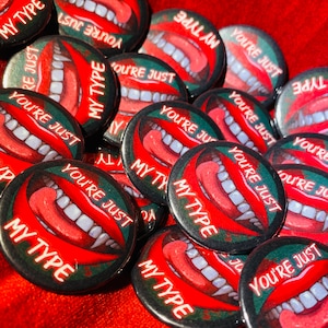 May include: A collection of round pins with a red, white and black design. The pins feature a graphic of a mouth with fangs and the text "My Type" and "You're Just".