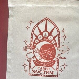 May include: A white canvas tote bag with a brown design of a crescent moon, stars, and clouds. The design is framed by a decorative arch and the text "CARPE NOCTEM" is printed below the design.