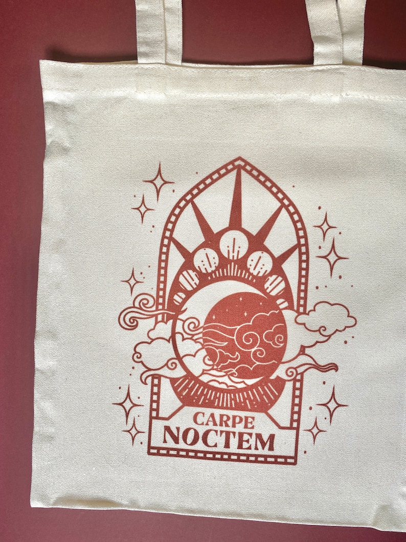 May include: A white canvas tote bag with a brown design of a crescent moon, stars, and clouds. The design is framed by a decorative arch and the text "CARPE NOCTEM" is printed below the design.