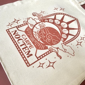 May include: A white canvas tote bag with a brown design of a crescent moon, stars, and clouds. The design is framed by a rectangle with the text "CARPE NOCTEM" inside.