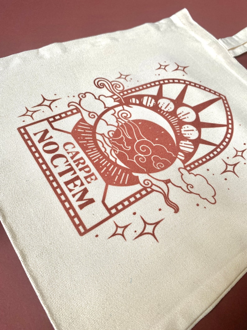 May include: A white canvas tote bag with a brown design of a crescent moon, stars, and clouds. The design is framed by a rectangle with the text "CARPE NOCTEM" inside.