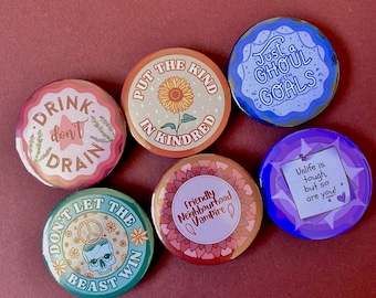 Peut inclure: Six boutons ronds colorés avec différents motifs et textes. Les boutons présentent un tournesol, un crâne, un vampire, un goule et un signe de paix. Le texte sur les boutons comprend "Drink don't drain", "Put the kind in kindred", "Just a ghoul with goals", "Friendly neighbourhood vampire", "Don't let the beast win", et "Unlife is tough, but so are you!"