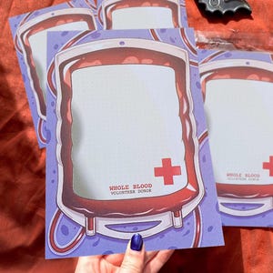 Könnte beinhalten: Mehrere Notizblöcke mit einem Blutbeutel-Design. Jeder Notizblock zeigt ein rotes Kreuz und den Text "WHOLE BLOOD VOLUNTEER DONOR". Eine kleine, silberne fledermausförmige Dekoration ist ebenfalls zu sehen.