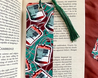 May include: A bookmark featuring a repeating pattern of red blood bags with white labels that say "Whole Blood" and a letter grade. The background is a green and red splatter pattern.