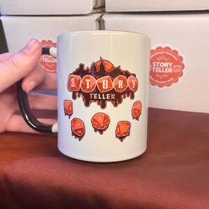 Peut inclure: Mug en c&eacute;ramique blanche avec une anse noire. Le mug pr&eacute;sente le texte "STORY TELLER" en rouge, ainsi que des illustrations de d&eacute;s rouges. Le mug a une finition brillante et est probablement destin&eacute; aux boissons chaudes.