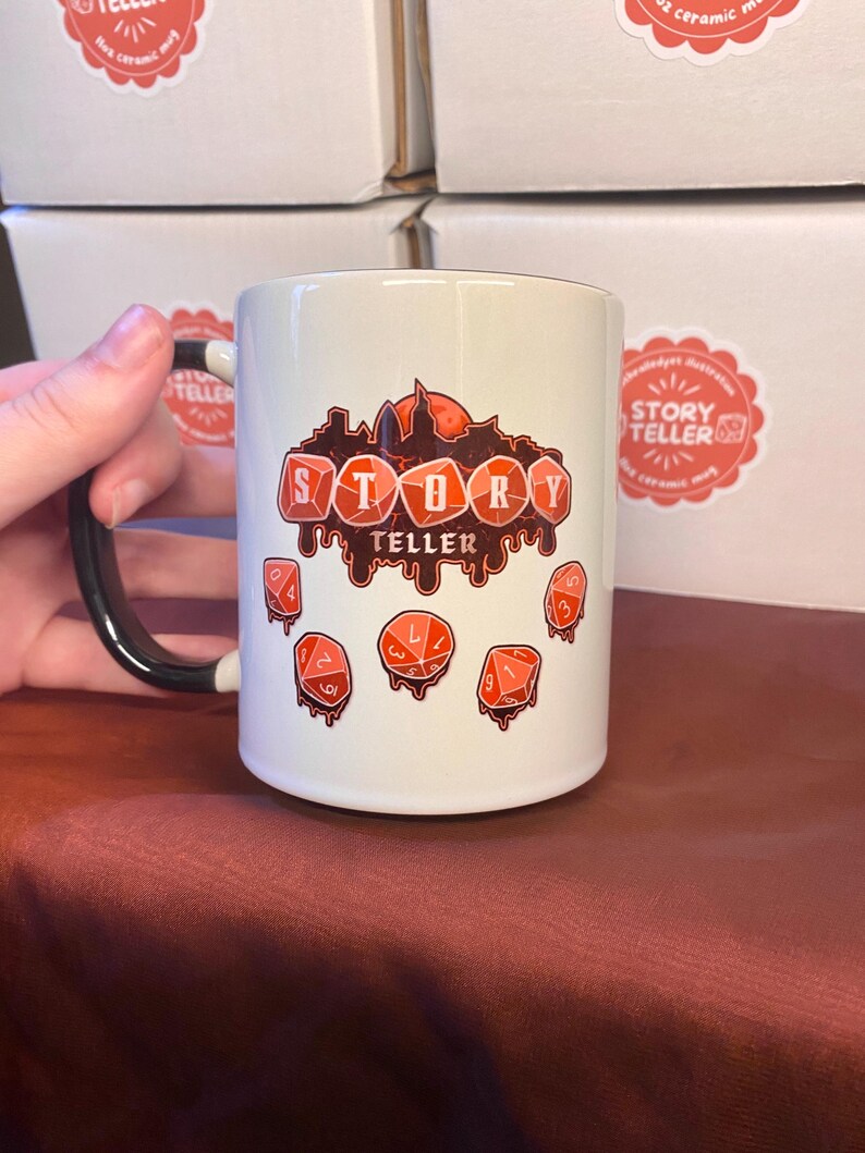 Peut inclure: Mug en c&eacute;ramique blanche avec une anse noire. Le mug pr&eacute;sente le texte "STORY TELLER" en rouge, ainsi que des illustrations de d&eacute;s rouges. Le mug a une finition brillante et est probablement destin&eacute; aux boissons chaudes.