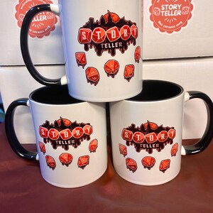 Peut inclure: Mugs en c&eacute;ramique blanches avec des anses et des bords noirs. Chaque mug porte l'inscription "STORY TELLER" en rouge d&eacute;goulinant, avec des illustrations de d&eacute;s rouges. Les mugs sont empil&eacute;s sur une surface marron.