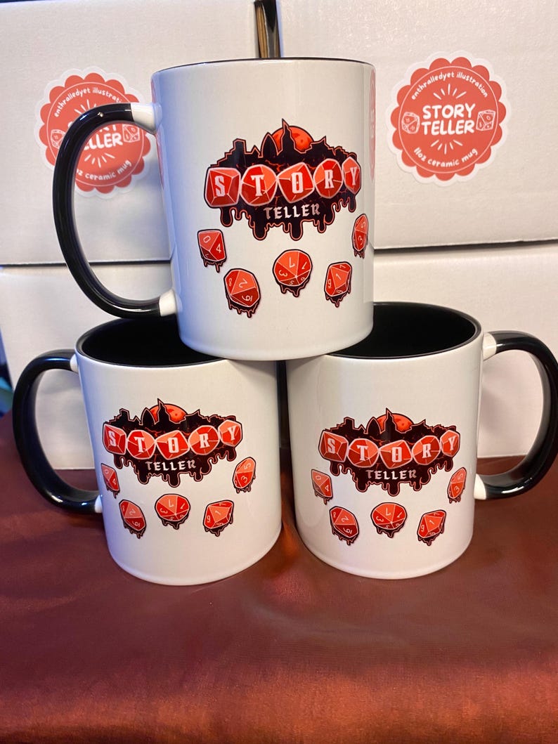 Peut inclure: Mugs en c&eacute;ramique blanches avec des anses et des bords noirs. Chaque mug porte l'inscription "STORY TELLER" en rouge d&eacute;goulinant, avec des illustrations de d&eacute;s rouges. Les mugs sont empil&eacute;s sur une surface marron.