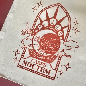 May include: A white tote bag with a brown design of a crescent moon, stars, and clouds. The design is framed by a rectangle with the text "CARPE NOCTEM" inside.