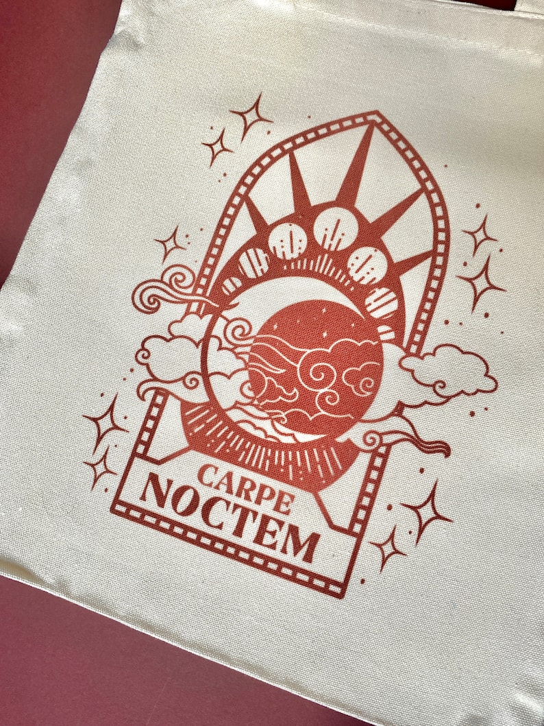 May include: A white tote bag with a brown design of a crescent moon, stars, and clouds. The design is framed by a rectangle with the text "CARPE NOCTEM" inside.