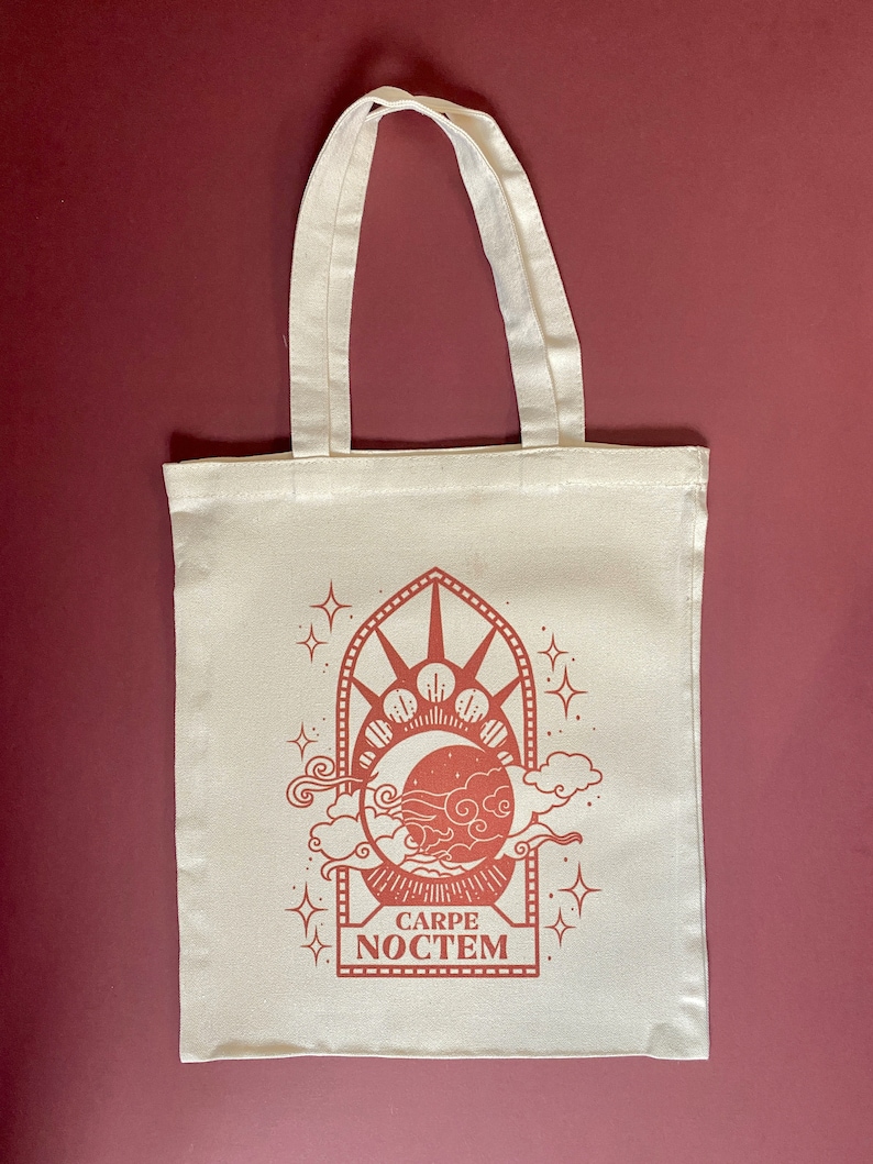 May include: A white tote bag with a red design featuring a crescent moon, stars, and clouds in a window frame. The text "CARPE NOCTEM" is printed below the design.