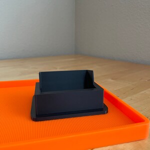 Puede incluir: Un soporte para teléfono de plástico negro con un borde elevado se encuentra sobre una alfombra de silicona naranja.