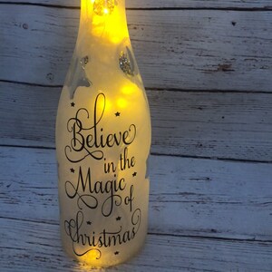 May include: A frosted glass bottle with a cork top and a string of lights inside. The bottle has a black vinyl decal that reads "Believe in the Magic of Christmas".