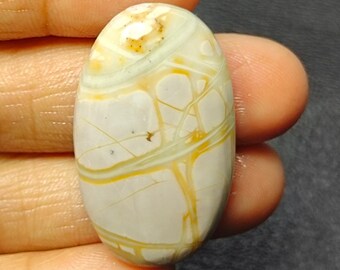 May include: An oval-shaped gemstone with a cream-colored base and intricate patterns of yellow and light green veins. The polished stone is held in a hand, showcasing its natural beauty and unique design.
