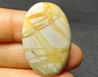 May include: Oval-shaped gemstone with a cream-colored base, crisscrossed by golden-yellow and pale green veins. The polished stone has a smooth surface and is held in a person's hand against a dark background. The gemstone is a natural stone.