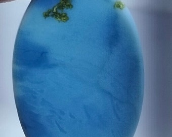 May include: A smooth, oval-shaped blue gemstone with green inclusions. The stone has a light blue color with white streaks throughout.