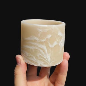 Puede incluir: Un portavelas cilíndrico de color beige cremoso con vetas blancas. El portavelas se sostiene en una mano sobre un fondo negro. Este artículo decorativo es un portavelas.
