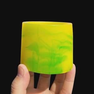 Puede incluir: Un vaso cilíndrico con un vibrante diseño veteado amarillo y verde. El material translúcido permite el paso de la luz, creando un efecto luminoso. El vaso se sostiene en una mano sobre un fondo negro.